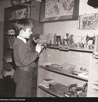chłopiec w szkolnym fartuchu stoi przy półce z zabawkami